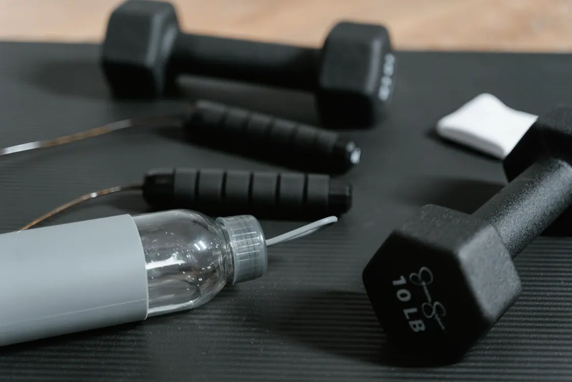 Black dumbbells, a jump rope and a clear water bottle on an exercise mat.